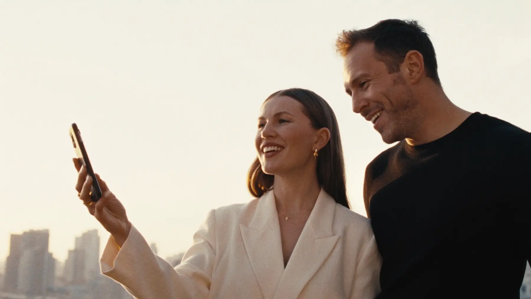 Two people take a phone video together on a rooftop at sunset