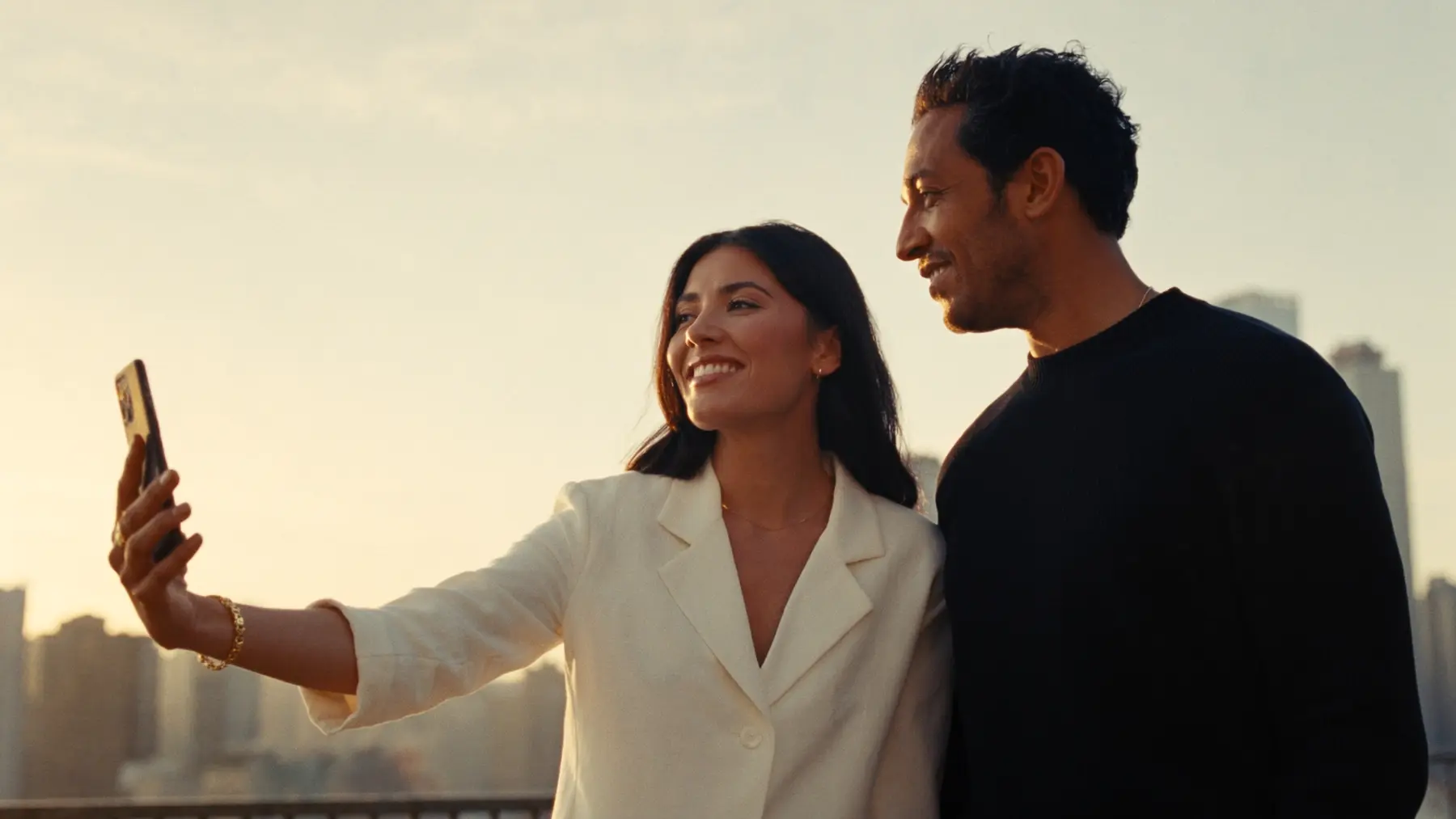 Two people take a phone video together on a rooftop at sunset