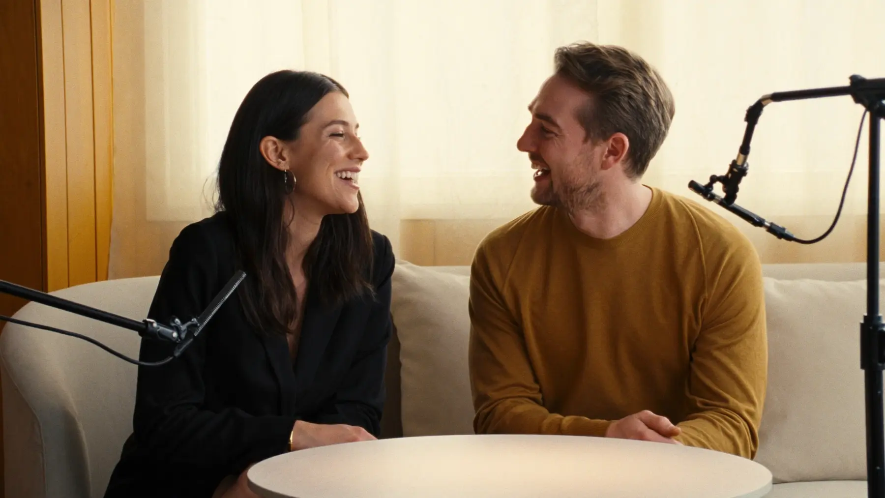 Two creators laugh together on a sofa with microphones, recording a conversation