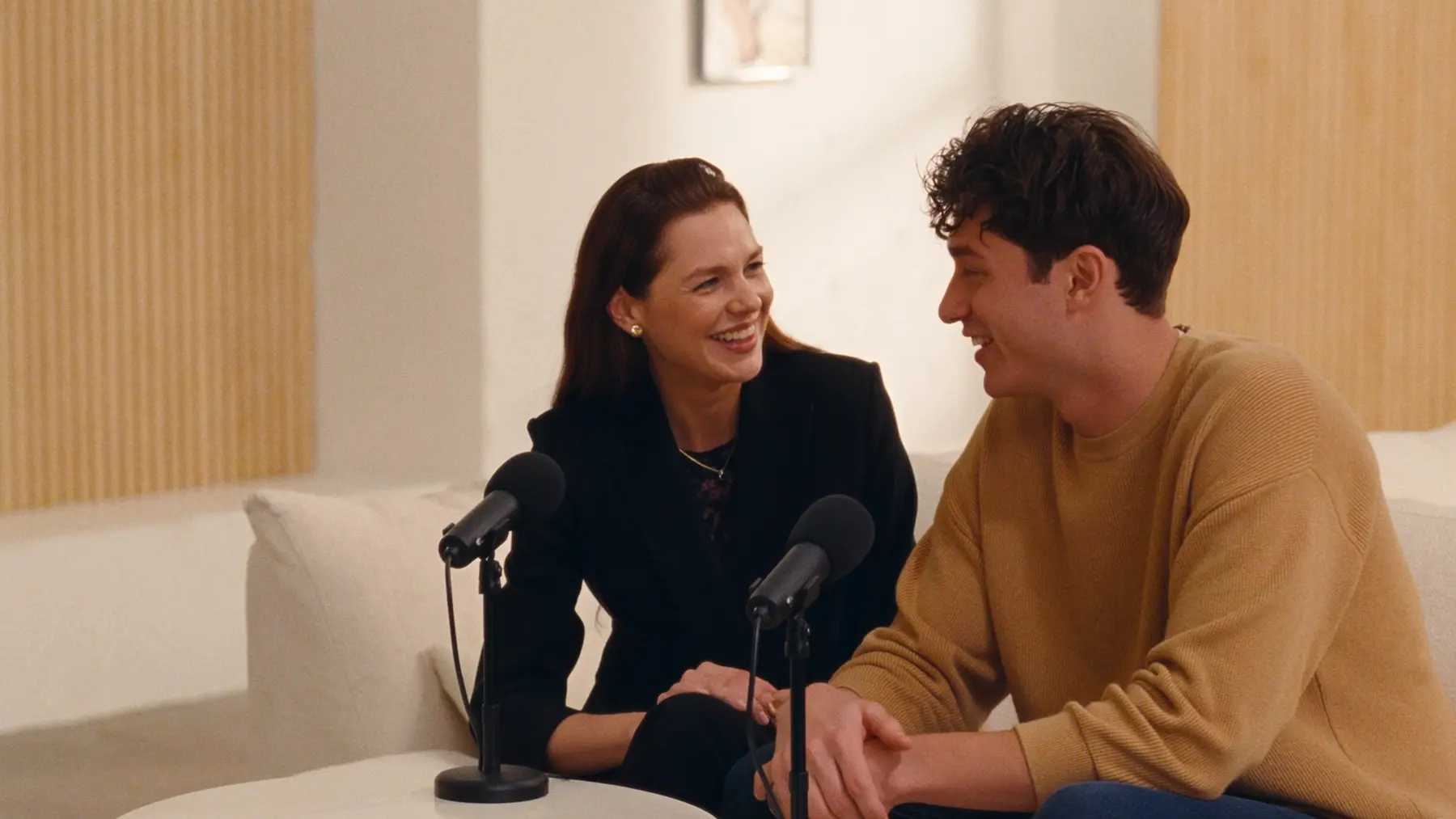Two creators laugh together on a sofa with microphones, recording a conversation