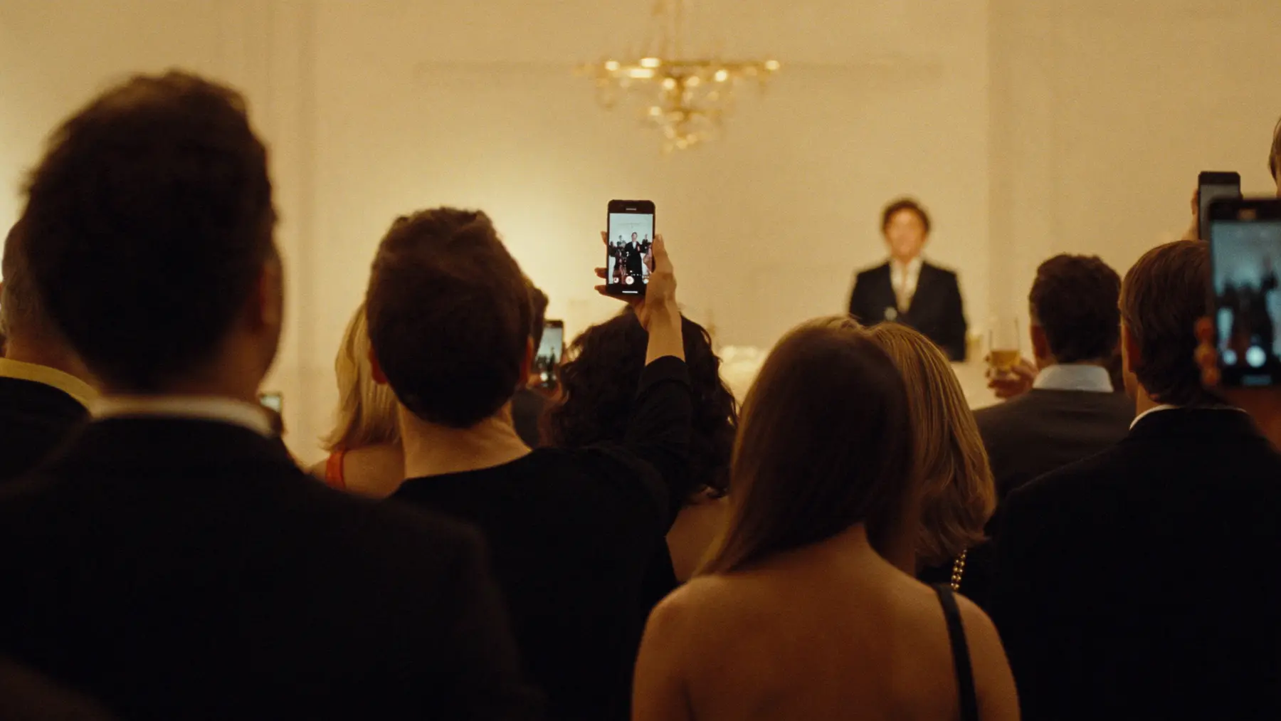 An audience in a grand room films a speaker on their phones
